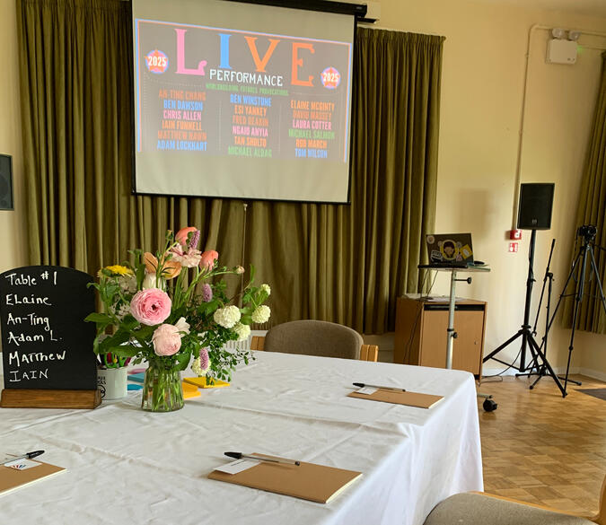a photo of a workshop room at Hawkwood College. There's a table with a white table cloth and notepads, pens and flowers, and a presentation screen in the background showing a Glastonbury festival style speaker lineup