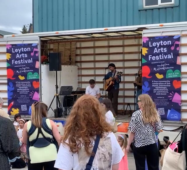a musician playing on a small stage built on the back of a lorry, with people watching. There are two large blue banners either side of the stage reading 'Leyton Arts Festival'